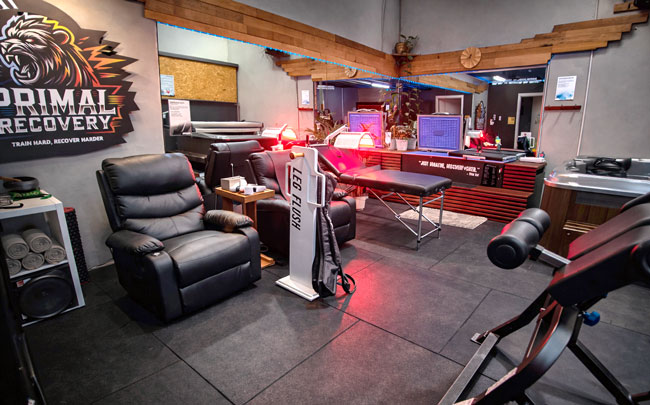 Industrial recovery space with ice baths, red light therapy, compression boots, massage table, inversion bench, and relaxation chairs arranged in a clean, dimly lit performance-focused environment.